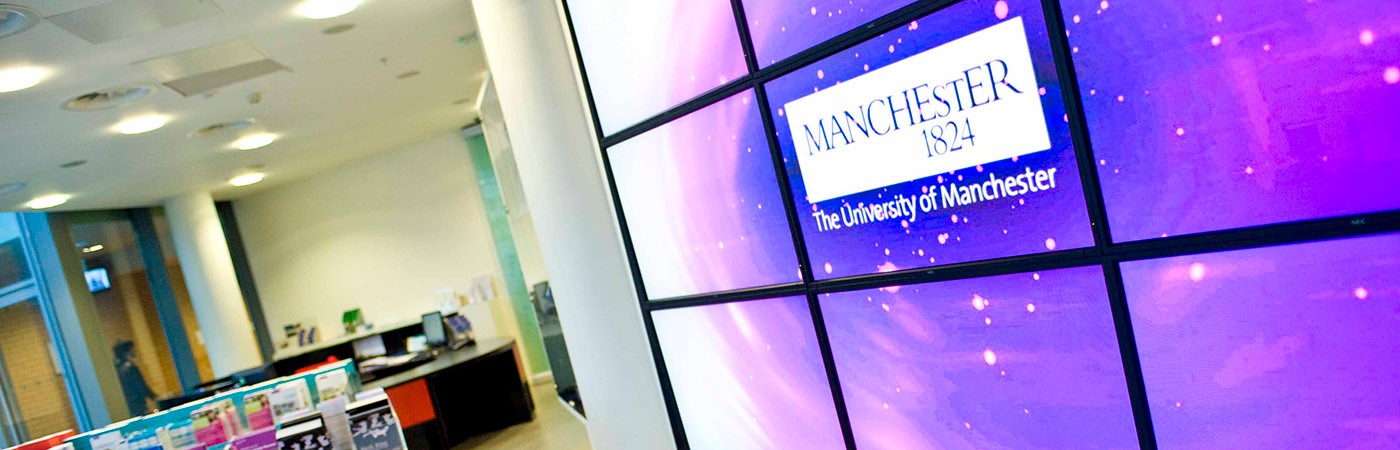 Digital display wall showing the words "Manchester 1824 – The University of Manchester" in a modern building interior with desks, brochures, and ceiling lights.